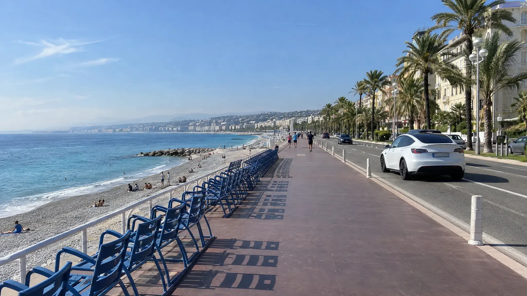 Tesla Model Y blanche garée le long de la Promenade des Anglais à Nice, les chaises bleues et la mer derrière