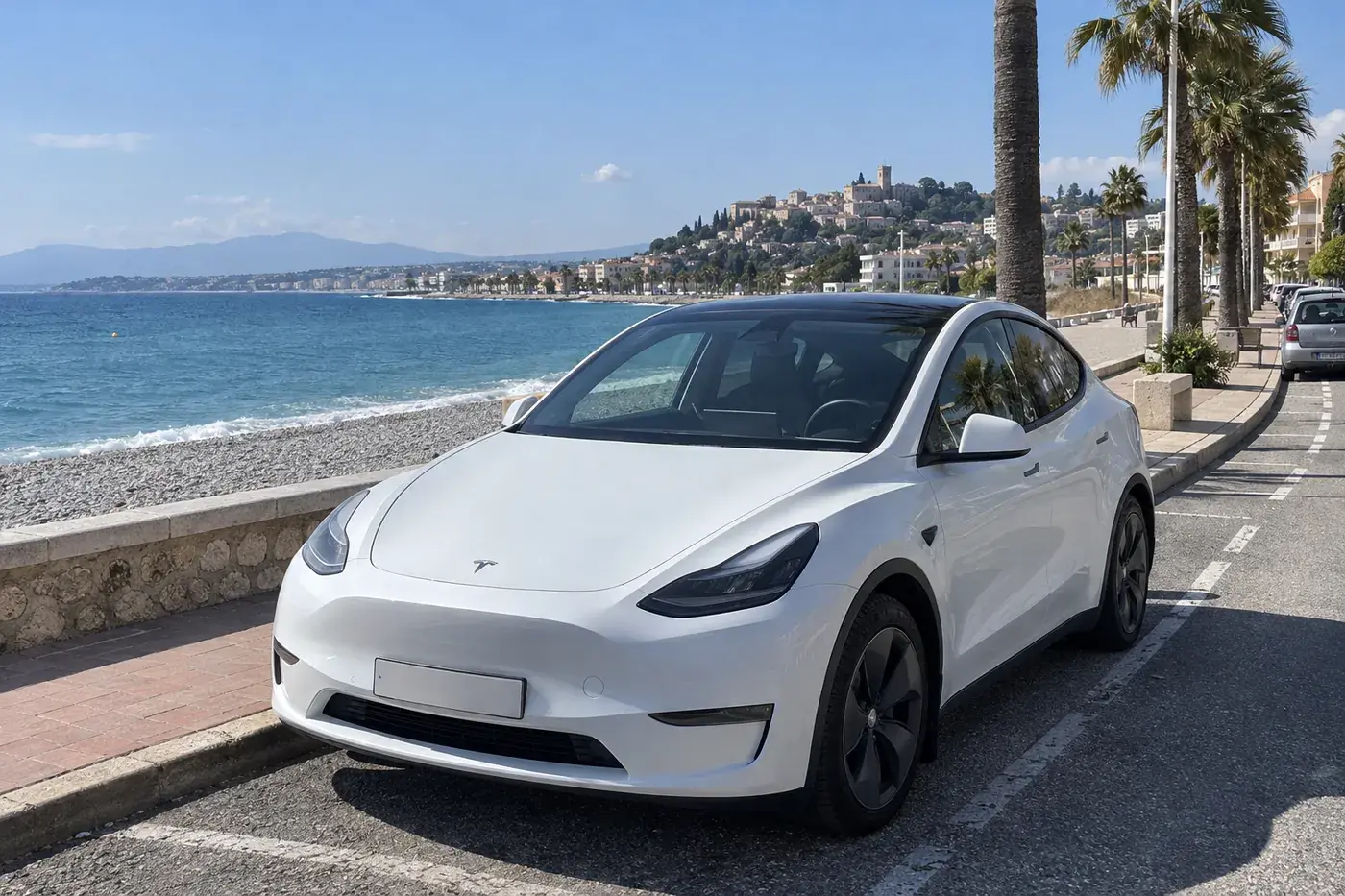 Tesla Model Y blanche garée sur le front de mer de Cagnes-sur-Mer, la plage de galets d’un côté, le Haut-de-Cagnes au loin