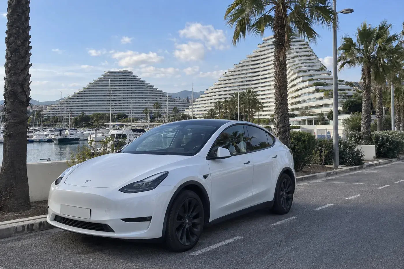 Tesla Model Y blanche garée près de la Marina Baie des Anges à Villeneuve-Loubet, les immeubles en vague et les palmiers derrière
