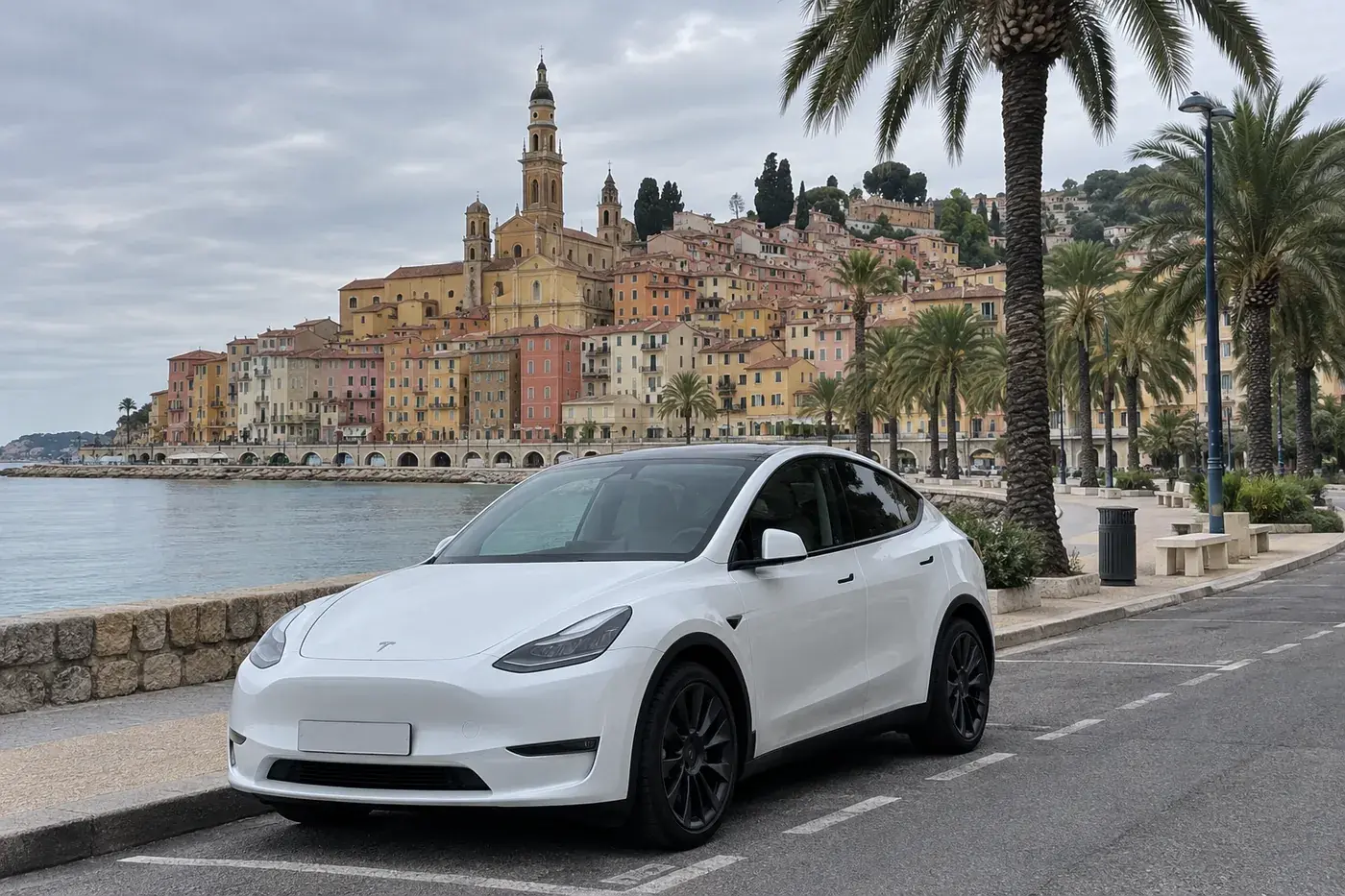 Tesla Model Y blanche garée sur la promenade du bord de mer à Menton, la vieille ville colorée et le clocher derrière