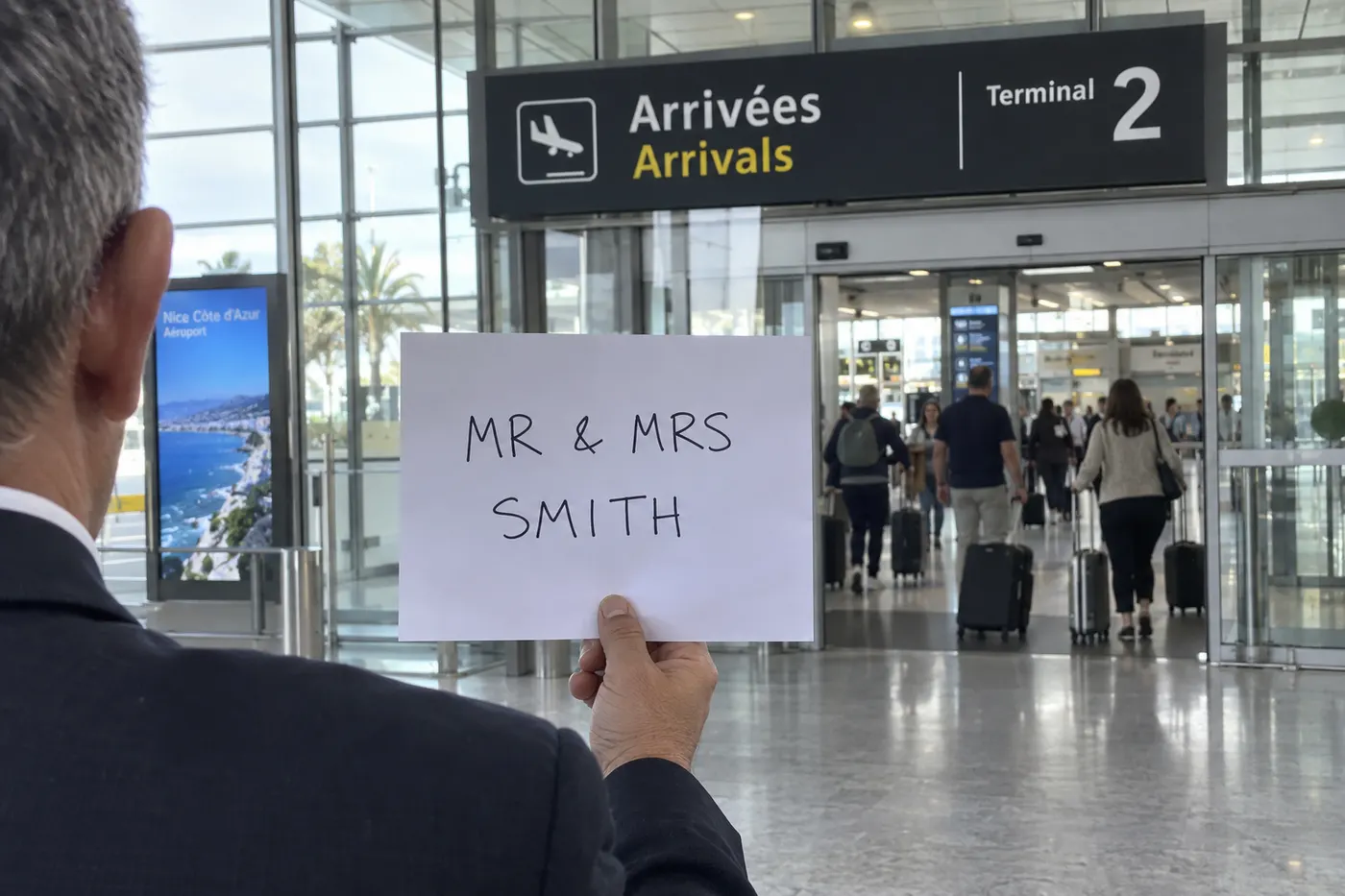 Une pancarte au nom de voyageurs tenue dans le hall des arrivées du Terminal 2 de l’aéroport de Nice, vue de dos